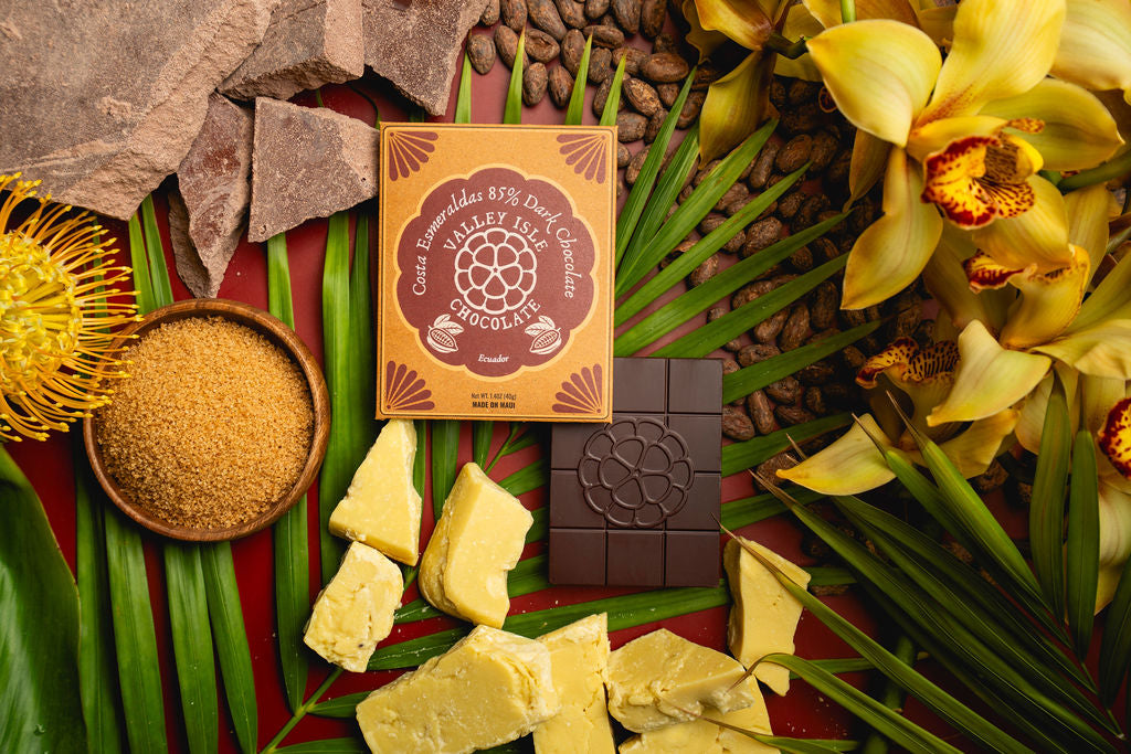 Chocolate bar with packaging surrounded by cocoa beans, chocolate pieces, and flowers on a textured surface.