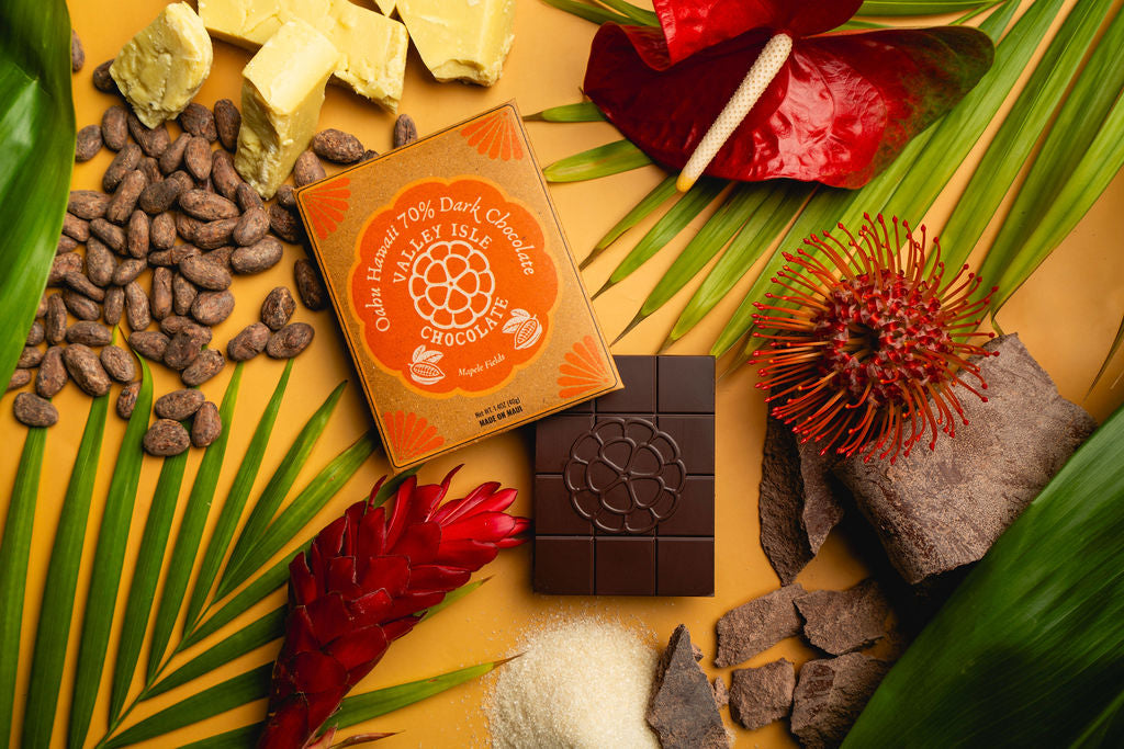 Dark chocolate bar with a box on a yellow surface with tropical leaves and flowers.