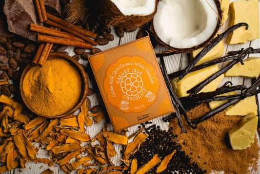 Box of Milky Isle Chocolates surrounded by ingredients like coconuts, vanilla beans, and turmeric on a wooden surface.